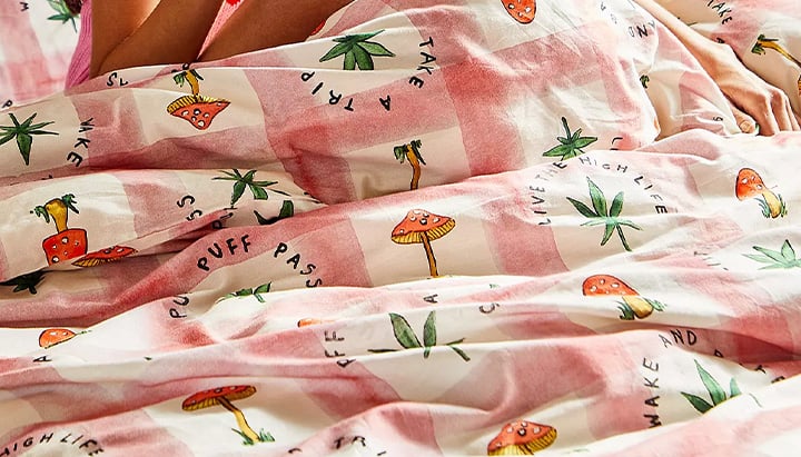 A person lying in bed under a pink and white checkered duvet cover with mushroom and cannabis leaf patterns