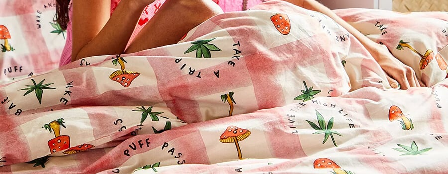 A person lying in bed under a pink and white checkered duvet cover with mushroom and cannabis leaf patterns