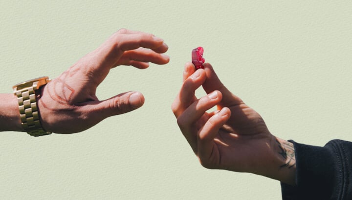 Two hands reaching towards each other, one with a tattoo, holding a red THC gummy bear.