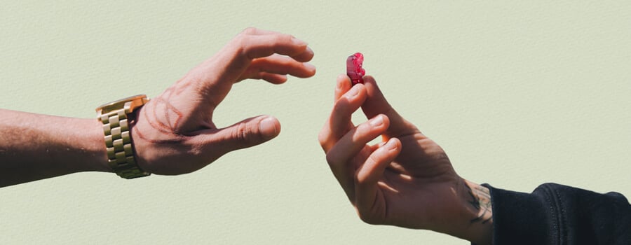 Two hands reaching towards each other, one with a tattoo, holding a red THC gummy bear.