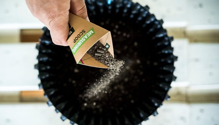 A hand holding a "MEASURING SCOOP" cardboard container, pouring a small amount of brown substance into a black pot for accurate measurement.