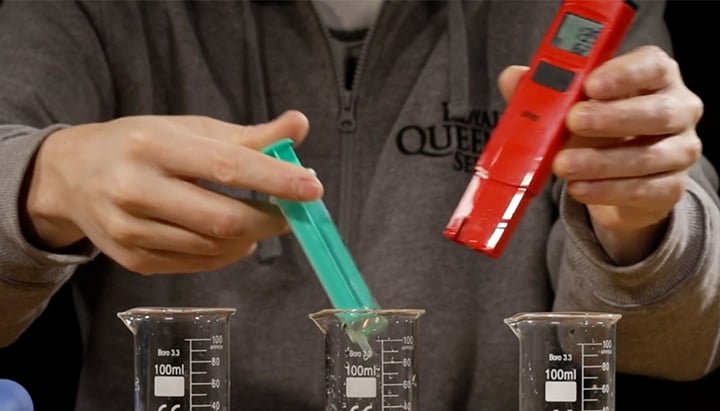 Person testing liquid pH using a pH meter and syringe. The syringe is about to be inserted into a beaker, with two other beakers visible on the table, for a pH measurement process.